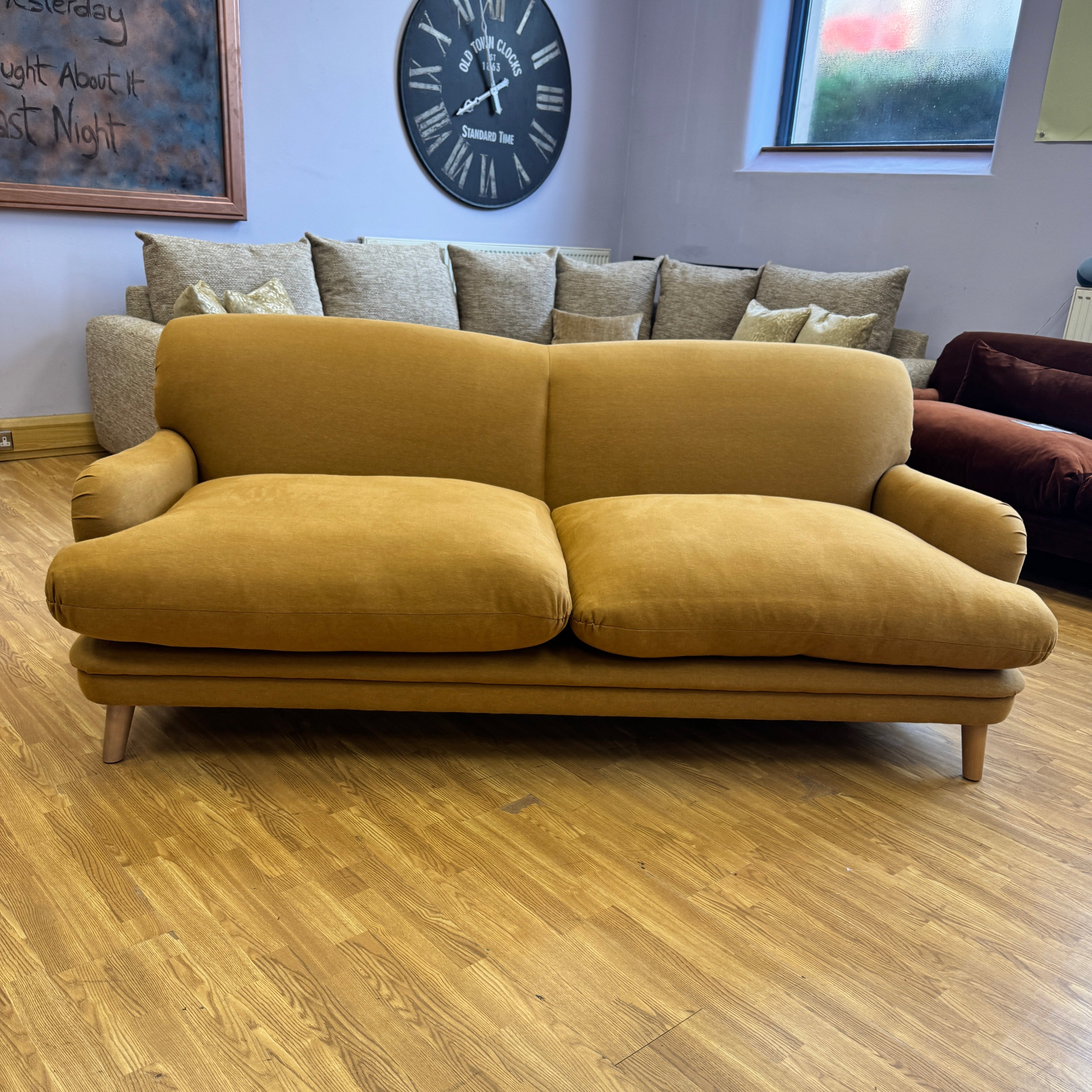 A mustard yellow fabric two-seater sofa with wooden legs, positioned on a light wood floor in a room with a window, a large wall clock, and other furniture in the background.